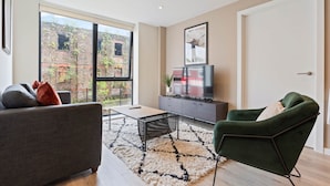 A modern living room with a sofa, armchair, TV, and a view of a brick building. - Liverpool Richmond Row 2 Bed/2 Bath Flat (107)| Secure Parking, WiFi & EV (Liverpool)