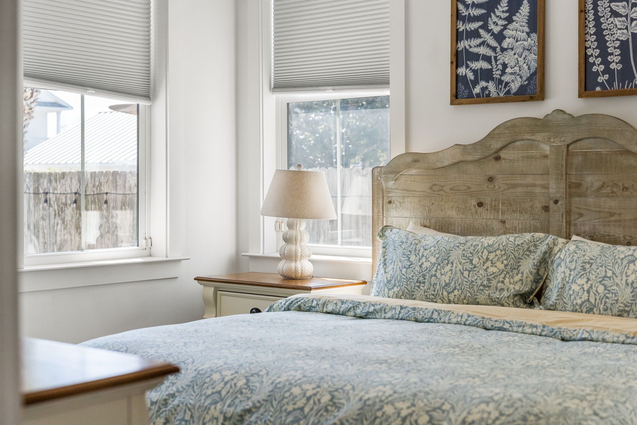 A bedroom with a wooden headboard, a bed with patterned bedding, a bedside lamp, and framed botanical artwork on the wall.