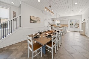 Uma sala de jantar espaçosa com uma mesa comprida posta para refeição, paredes brancas, uma escadaria e uma cozinha ao fundo.
