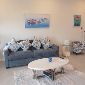 A living room with a grey sofa, patterned cushions, a round coffee table, a starfish rug, and two paintings on the wall. - New Oceanfront Beach House, "Paradise Blue" - Your Private Tropical Escape. (Rolleville)