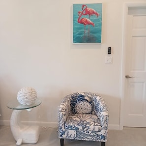 A room with a white door, a glass table with a white sculpture, a patterned armchair, and a painting of flamingos.