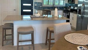 A modern kitchen with a central island, bar stools, and a dining table. - New Oceanfront Beach House, "Paradise Blue" - Your Private Tropical Escape. (Rolleville)