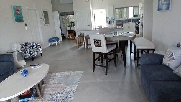 A spacious living area with a dining table, chairs, a sofa, and a kitchen in the background. - New Oceanfront Beach House, "Paradise Blue" - Your Private Tropical Escape. (Rolleville)