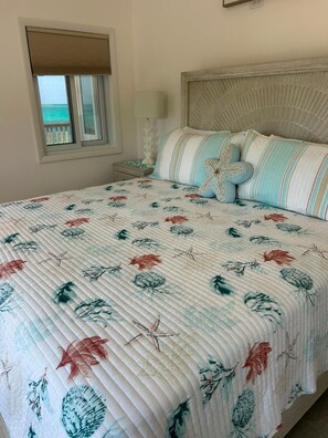 A bed with a floral quilt, a window with a view, and a bedside lamp. - New Oceanfront Beach House, "Paradise Blue" - Your Private Tropical Escape. (Rolleville)
