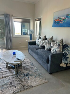 Un salon avec un canapé gris, des coussins à motifs, une table basse ronde sur laquelle se trouve un objet en verre bleu, une lampe bleue et un tableau accroché au mur.