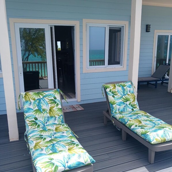 A covered outdoor area with two lounge chairs, a sliding glass door, and a view of the ocean.