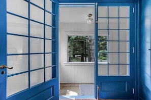 A blue door with glass panels, a window with a view of trees, and a light fixture above the door. - Reef Point Cottage-Ocean views! (Bar Harbor)