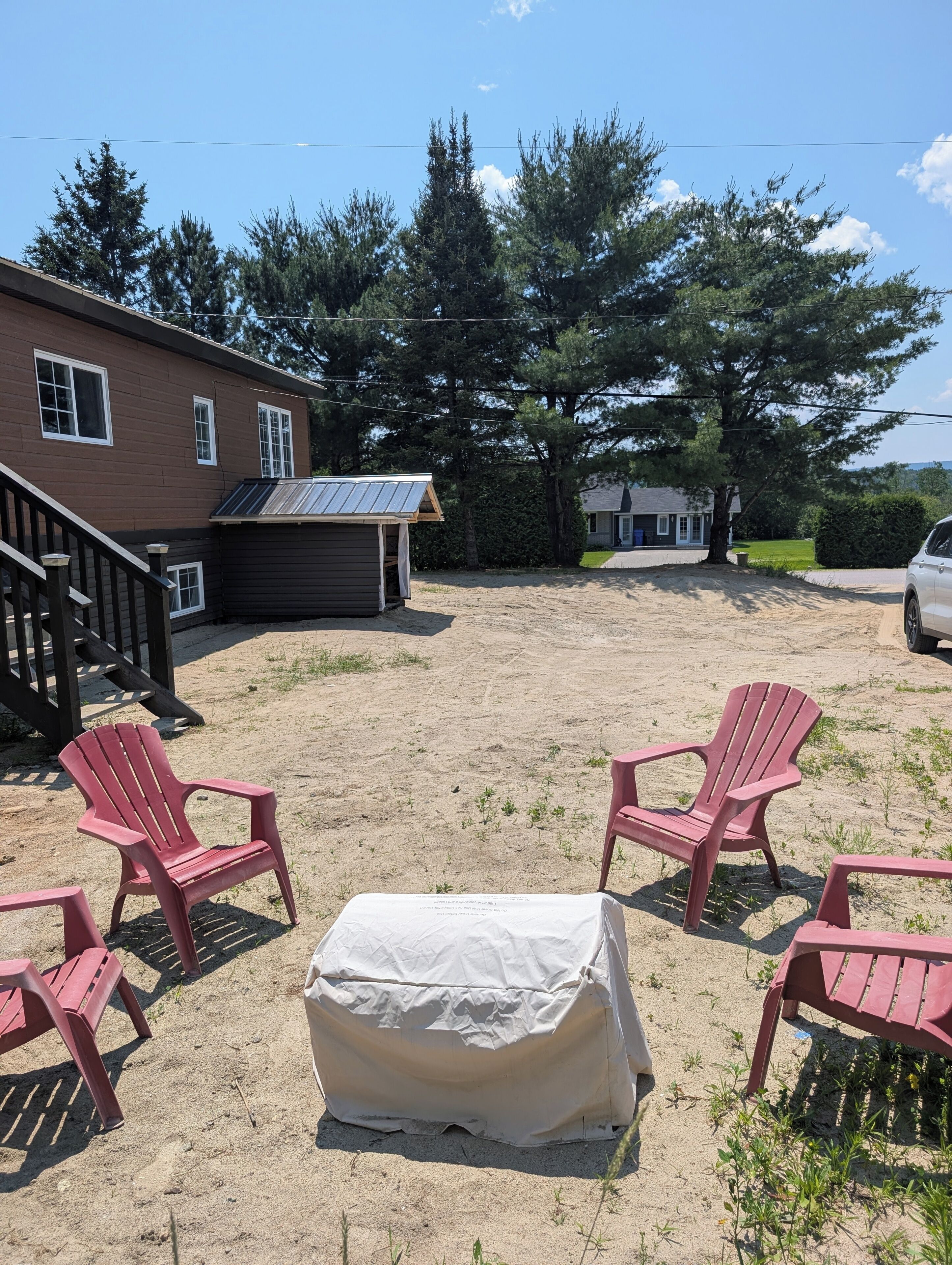 A backyard with a covered picnic table, red Adirondack chairs, a wooden deck, and a house with a metal roof.