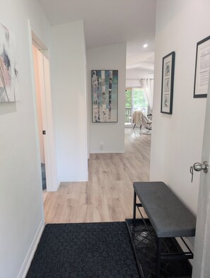 A hallway with a bench, artwork on the walls, and a view into a room with a chair.