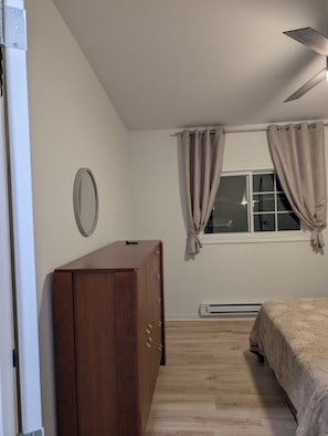 A bedroom with a wooden wardrobe, a bed with a floral bedspread, a window with curtains, and a ceiling fan.