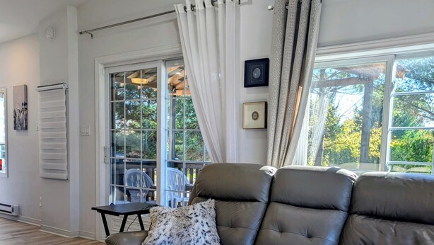 A modern living room with a leather sofa, a small table, and a chair. There is a window with curtains, a ceiling fan, and a view of trees outside.