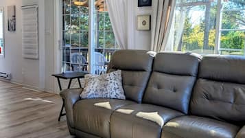 A modern living room with a leather sofa, a small table, and a chair. There is a window with curtains, a ceiling fan, and a view of trees outside.