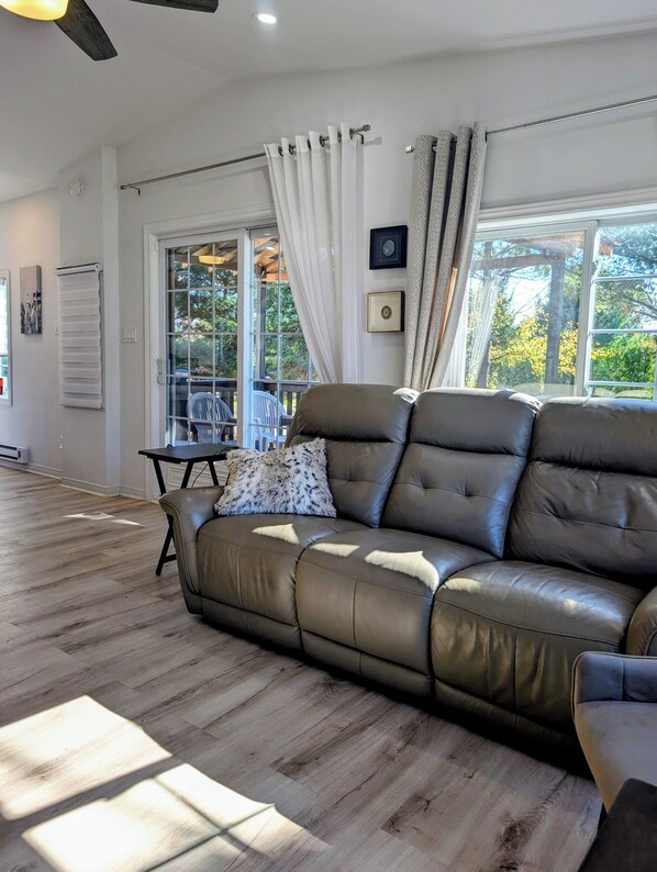 A modern living room with a leather sofa, a small table, and a chair. There is a window with curtains, a ceiling fan, and a view of trees outside.