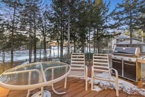 Een terras met een glazen tafel en metalen stoelen, een barbecue, en uitzicht op een besneeuwd landschap met bomen.