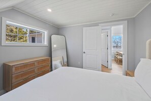 A bedroom with a large bed, a dresser, a mirror, and a door leading to a dining area. - The Cove on Newbury Neck (Surry)