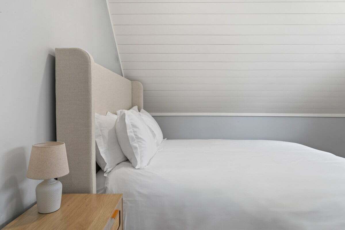 Una cama bien tendida con sábanas blancas, una lámpara de noche y un cabecero frente a una pared lisa.