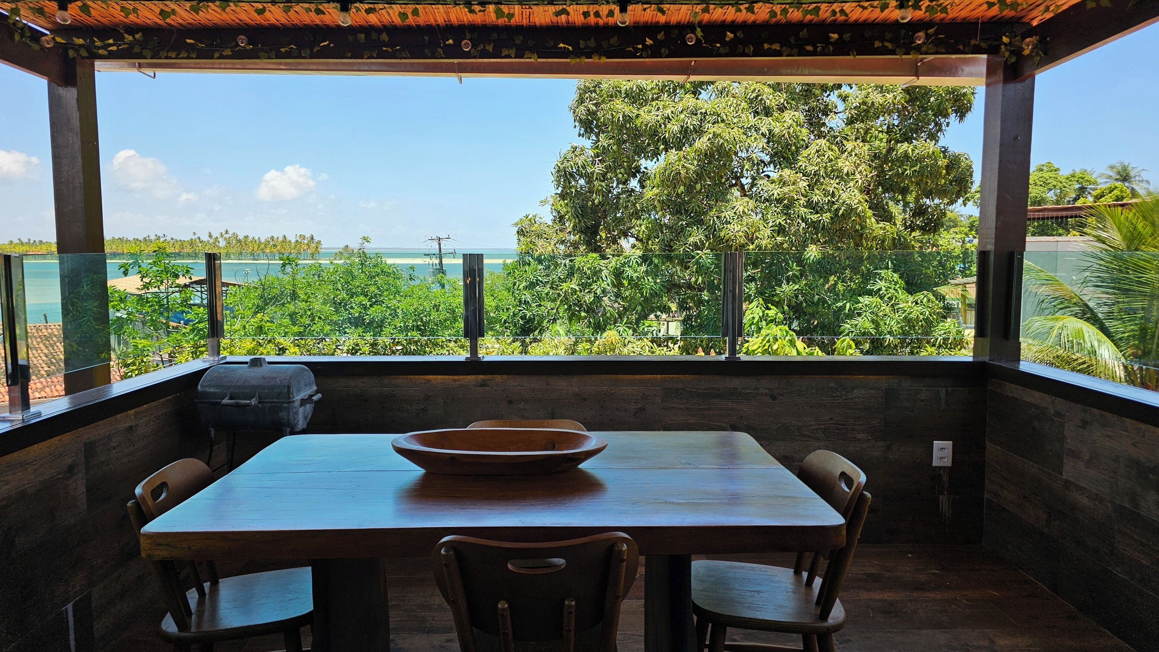 A covered outdoor dining area with a table and chairs, overlooking a scenic view of trees and a body of water.
