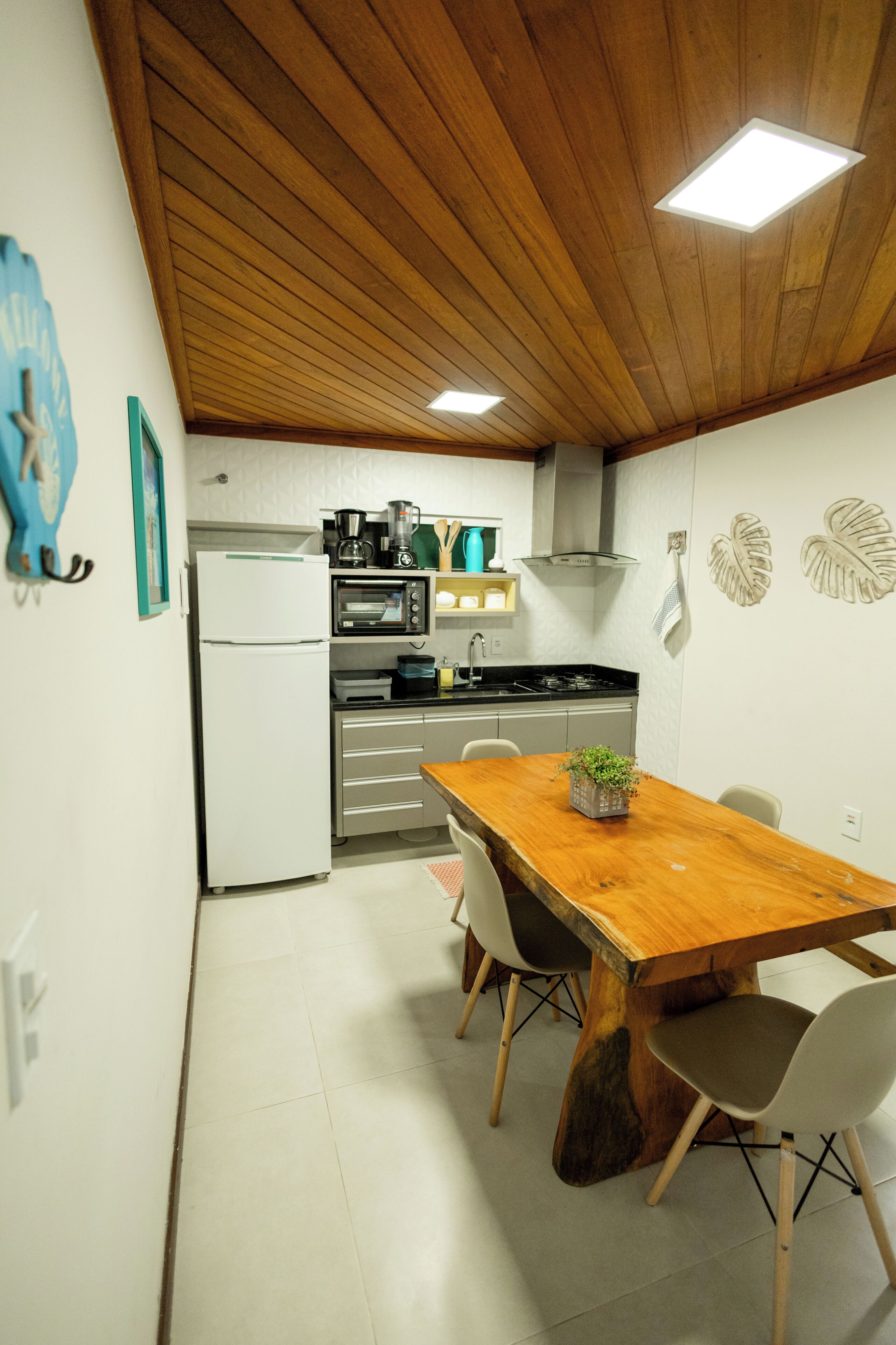 Une cuisine compacte équipée d’un réfrigérateur, d’un four et d’une machine à café, une table à manger en bois avec des chaises, et un plafond doté d’un éclairage encastré.