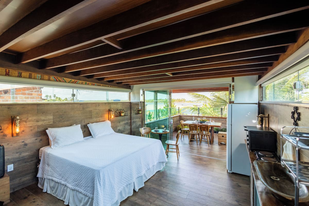 A bedroom with a bed, wooden ceiling beams, a dining area with chairs and a table, and a kitchen area with a sink and stove.