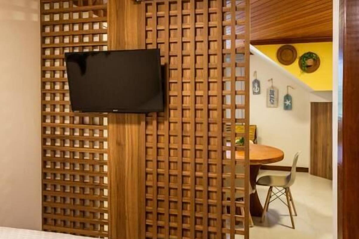 A bedroom with a bed, a wooden partition, a television mounted on the wall, and a dining area visible through an open door.