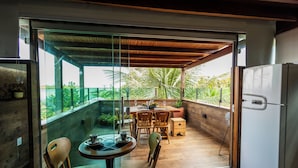 A modern kitchen with a refrigerator, wooden chairs, a dining table, and a glass door leading to a balcony with a view of palm trees. - Pousada Feliz Boipeba Apartamentos (Cairu)