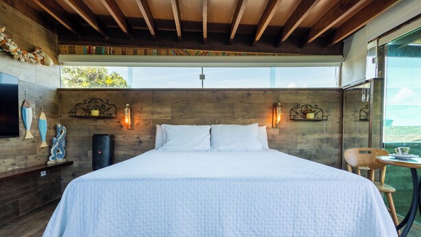 A large bed with a white quilt, a wooden ceiling, a wall-mounted TV, and a window overlooking a landscape. - Pousada Feliz Boipeba Apartamentos (Cairu)
