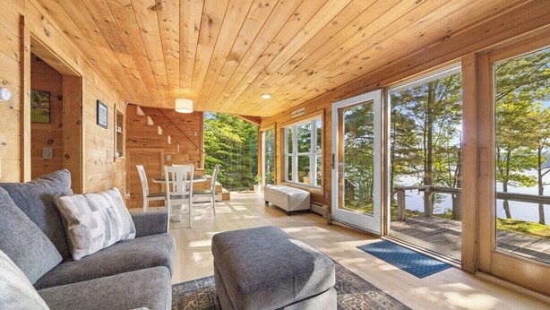 A cabin interior with a wooden ceiling, a sofa, a bench, and a dining area with a view of the outdoors.