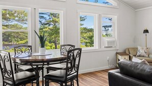 A room with a dining table, chairs, a sofa, and large windows with a view of trees. - Shoreline Solace - Waterfront! Close to Acadia! (Ellsworth)