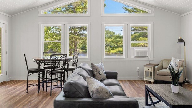 A spacious room with a high ceiling, large windows, a dark leather sofa, a dining table with chairs, and a small side table. - Shoreline Solace - Waterfront! Close to Acadia! (Ellsworth)