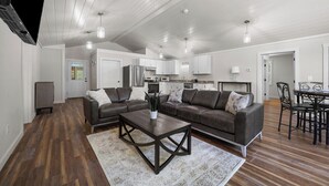 A spacious living room with a dark wood floor, a large L-shaped sofa, a coffee table, and a dining area with a table and chairs. - Shoreline Solace - Waterfront! Close to Acadia! (Ellsworth)