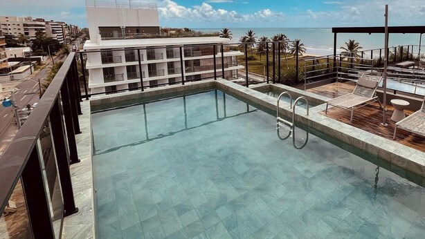 Una piscina en la azotea con agua azul cristalina, rodeada por un edificio moderno con balcones y vistas al océano.