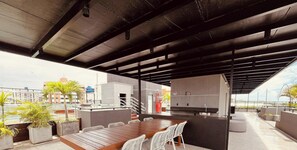 A rooftop terrace with a wooden table, white chairs, potted plants, a kitchen area, and a view of the cityscape. - LM SUN INTERMARES (Cabedelo)
