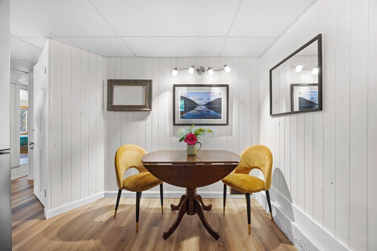 A dining area with a round wooden table, two yellow chairs, a vase with flowers, and framed pictures on the walls.