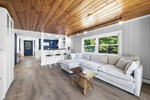 A modern living room with a wooden ceiling, a large sofa, a coffee table, and a kitchen area in the background. - Golden Hour Retreat - Hot Tub & Lake Views (Ellsworth)
