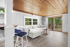 Una sala de estar moderna con techo de madera, paredes blancas, un sofá blanco, cojines azules y una mesa de madera.