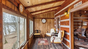 A wooden cabin interior with a large window, a clock, a dining area with chairs, and a sofa.