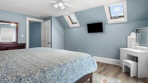 Une chambre à coucher comprenant un lit, un bureau, une chaise, un ventilateur de plafond et un téléviseur fixé au mur.