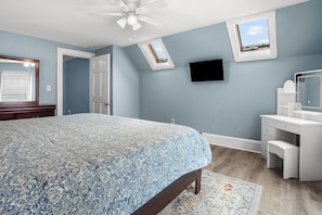 A bedroom with a bed, a desk, a chair, a ceiling fan, and a TV mounted on the wall.