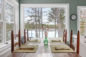 A dining table with a view of a lake and trees, wooden chairs, and a green vase with branches. - Tidal Falls - Waterfront! (Hancock)