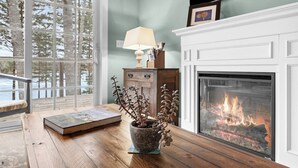 Une pièce avec une cheminée, une fenêtre donnant sur des arbres, une table avec un livre et une plante en pot, et une lampe.