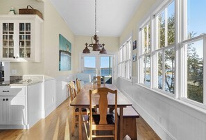 Uma sala de jantar com mesa e cadeiras de madeira, luminária pendente e grandes janelas com vista para o exterior.