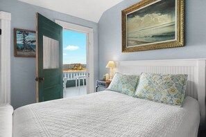 Quarto com cama, pintura, porta que dá para uma varanda e mesa de cabeceira com abajur.
