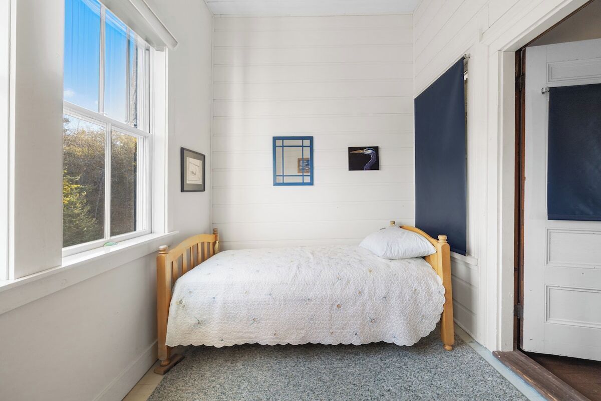 Ein kleines Zimmer mit einem Einzelbett, einem hölzernen Kopfteil, einem Fenster mit Jalousien, einer Tür und einem gerahmten Bild an der Wand.