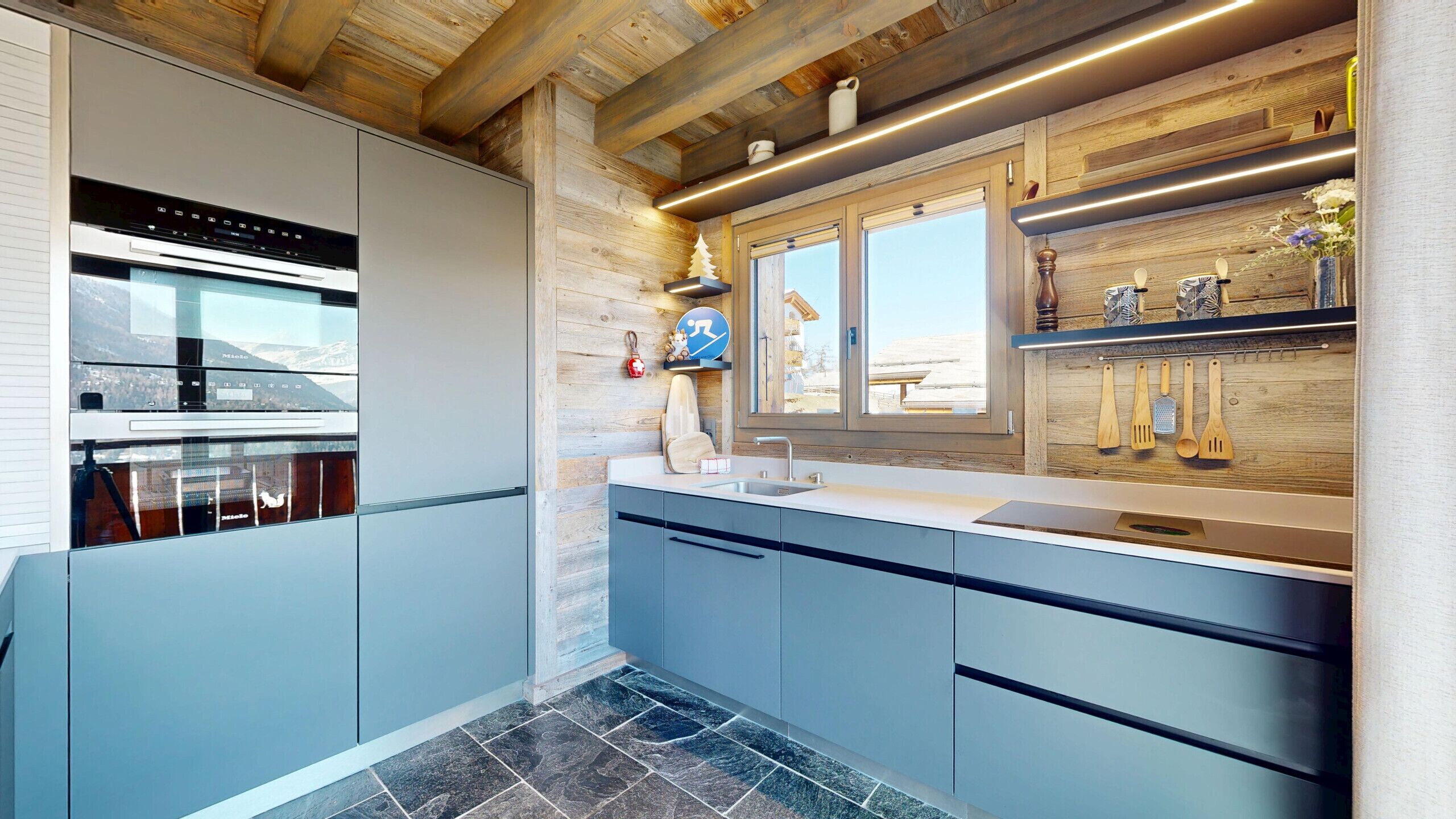 Una cuina moderna amb forn integrat, finestra amb vistes i prestatges de fusta amb utensilis de cuina.