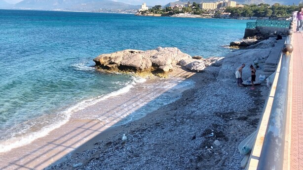 Eine Küstenpromenade mit Kiesstrand, einem Felsen und einem entfernten Gebirgszug.