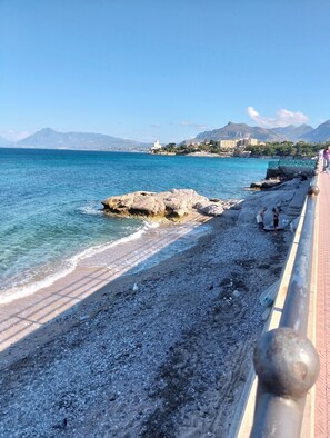 Eine Küstenpromenade mit Kiesstrand, einem Felsen und einem entfernten Gebirgszug.