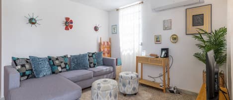 Ein Wohnzimmer mit grauem Sofa, gemusterten Kissen, zwei Hocker, einem Couchtisch aus Holz mit einem Fernseher und dekorativen Wanduhren.
