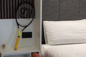 A bed with white bedding, a grey headboard, and a white nightstand with a tennis racket. - Luxury Westport Studio | Premium Shower + WiFi (Westport)