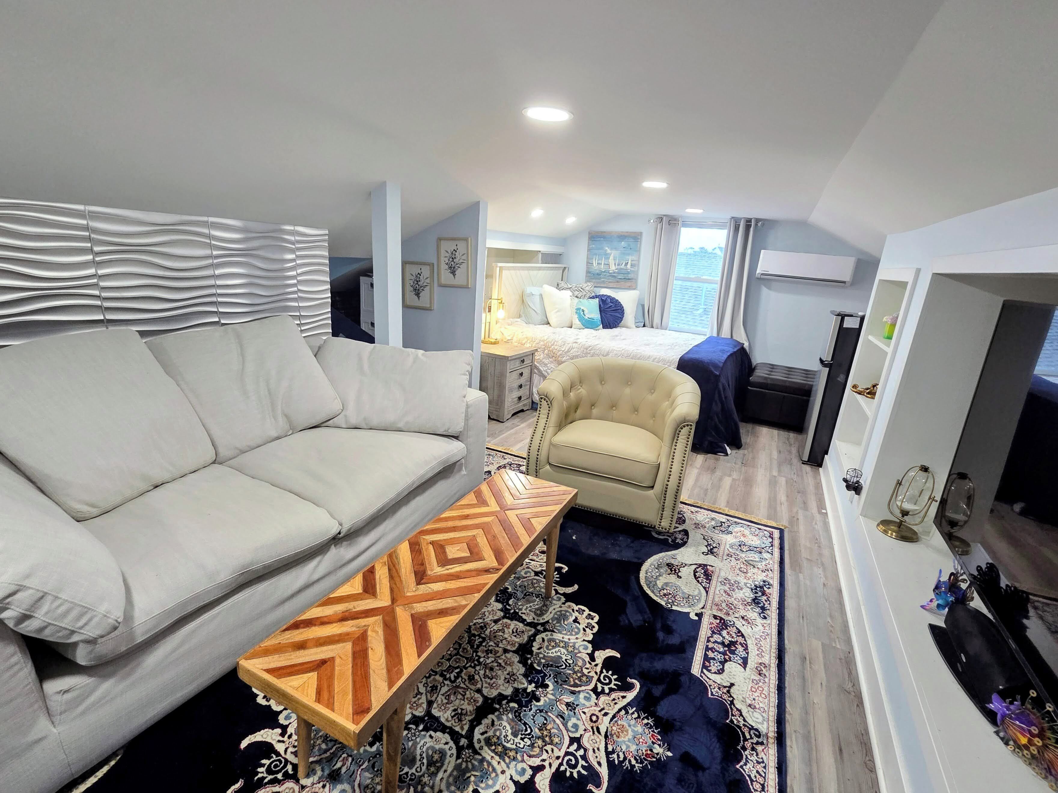 A modern hotel room with a sofa, armchair, patterned rug, and a bed with a view.