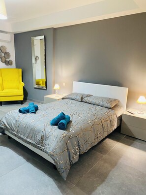 Een moderne slaapkamer met een grijs bed, twee bedlampjes, een gele fauteuil en een grote spiegel aan de muur.